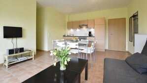 A living room with a TV, a coffee table with a vase of flowers, a grey sofa, and a kitchen area with cabinets and a dining table. - Quaint Apartment in Zierow near Sea (Zierow)