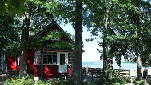 Een rode blokhut met een veranda, omgeven door bomen en uitzicht op het water.