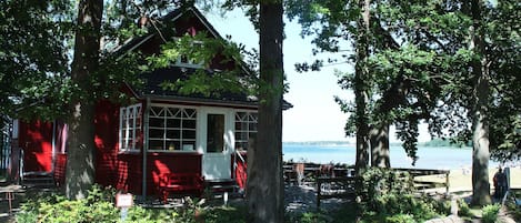 A red cabin with a porch, surrounded by trees and a view of the water.
