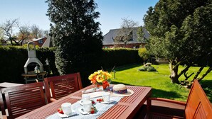 A wooden deck with a table set for two, surrounded by chairs, and a barbecue grill in the background.