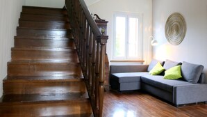 Una escalera de madera que da a un rellano, un sofá gris con cojines verdes, y una lámpara fijada a la pared.