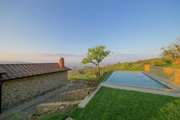 A stone building with a tiled roof, a swimming pool, and a view of a hilly landscape.