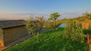 Un bâtiment en pierre avec un toit en tuiles, entouré de pelouse et d’arbres, sous un ciel dégagé.