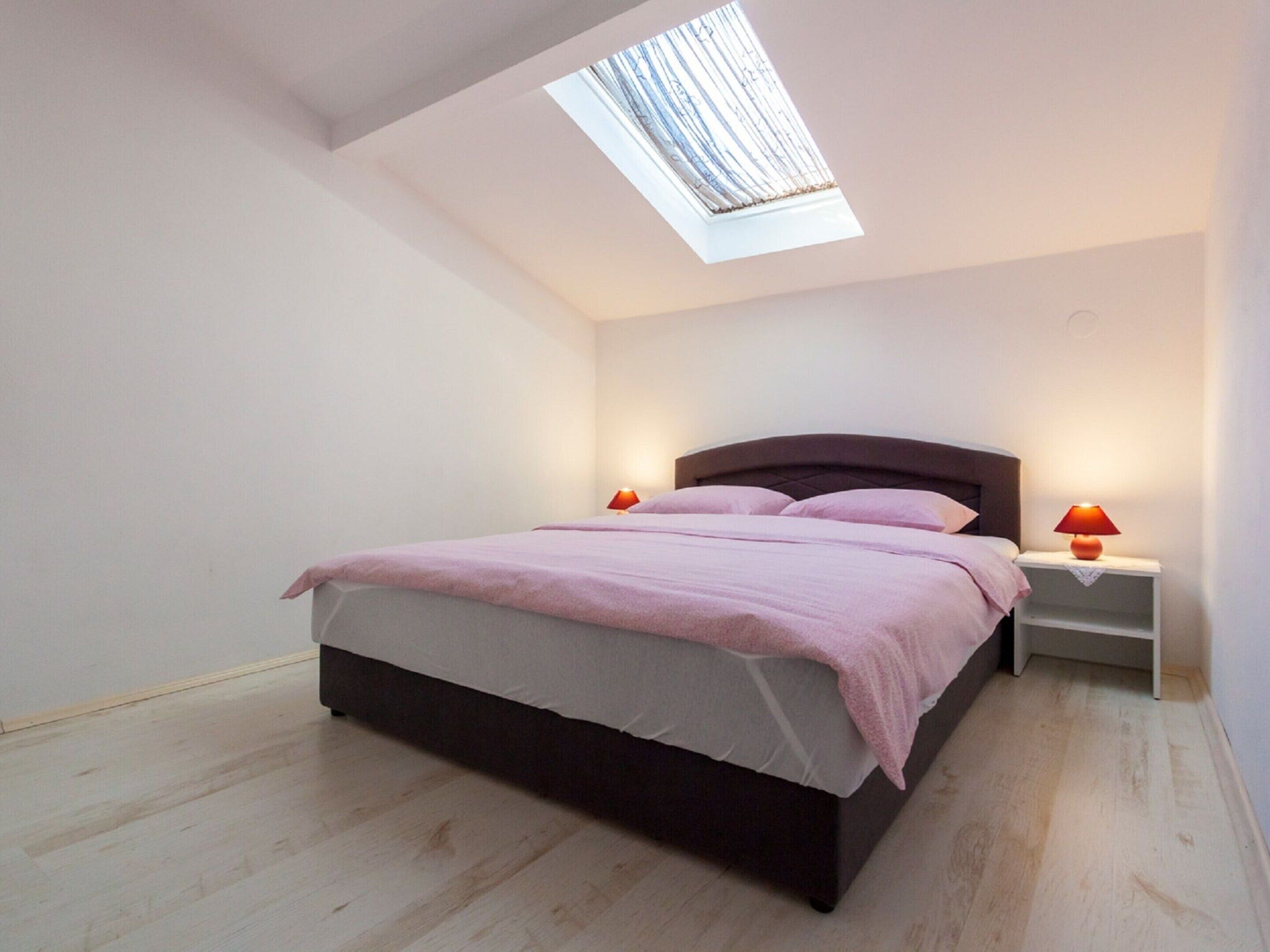 A bedroom with a skylight, a bed with pink bedding, two bedside tables with lamps, and a wooden floor.