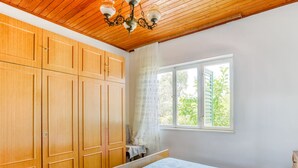 Une chambre avec des armoires en bois, un lit avec une literie à motifs, une fenêtre donnant sur un paysage verdoyant et un lustre.