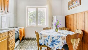 Un coin repas avec une table ronde recouverte d’une nappe bleue et blanche, des chaises en bois, une fenêtre donnant sur des arbres et un tableau accroché au mur.