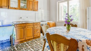 Une cuisine avec des meubles en bois, un pare-feu en carrelage, une table à manger recouverte d’une nappe à motifs floraux et une fenêtre avec des rideaux.