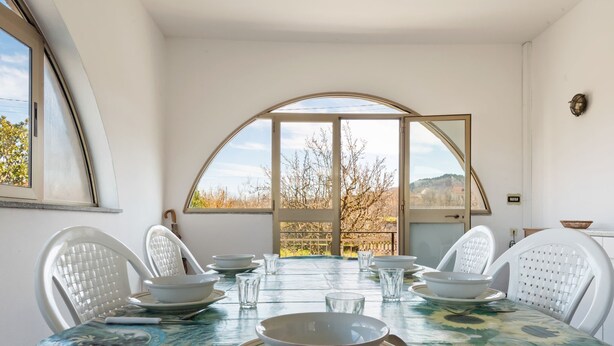A dining area with a table set for four, white chairs, and a view through large arched windows. - Apartment in Cassinelle With Garden & BBQ (Cassinelle)