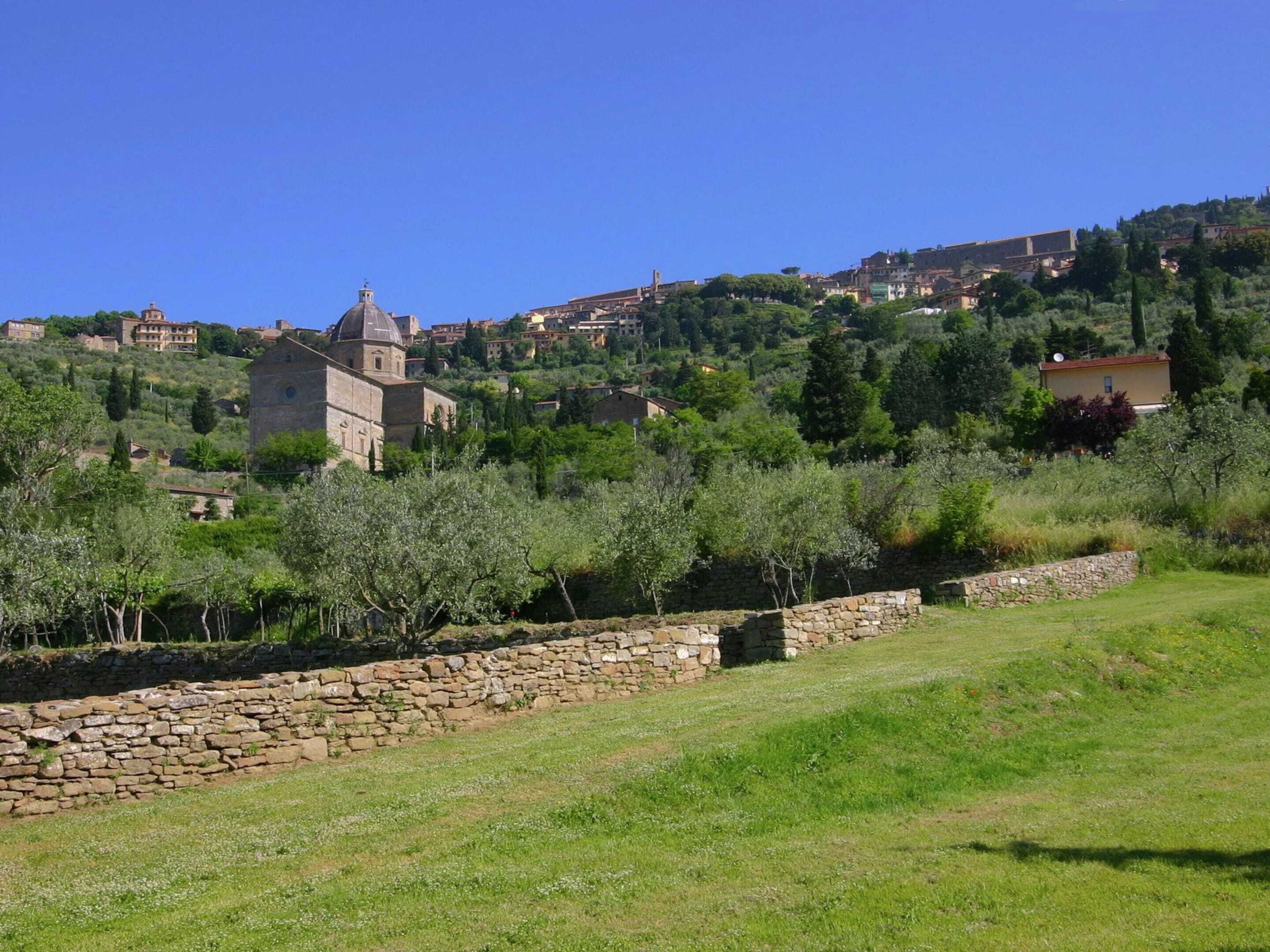 Un mur de pedra, un camp amb herba i una collada amb edificis.