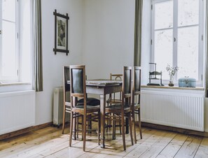 Un comedor con una mesa redonda y cuatro sillas, un radiador, una ventana con cortinas y un cuadro en la pared.
