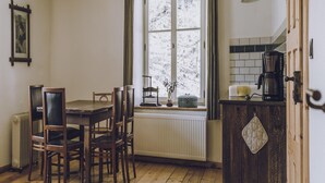 Una zona de menjador amb taula i cadires, una finestra amb cortines, un radiador i una cuina amb cafetera.
