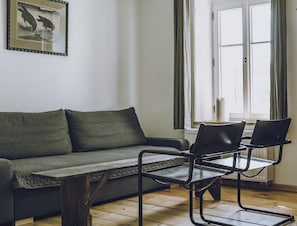 Una sala de estar con un sofá, una mesa de centro y dos sillones. Se ve una ventana con cortinas y un radiador.