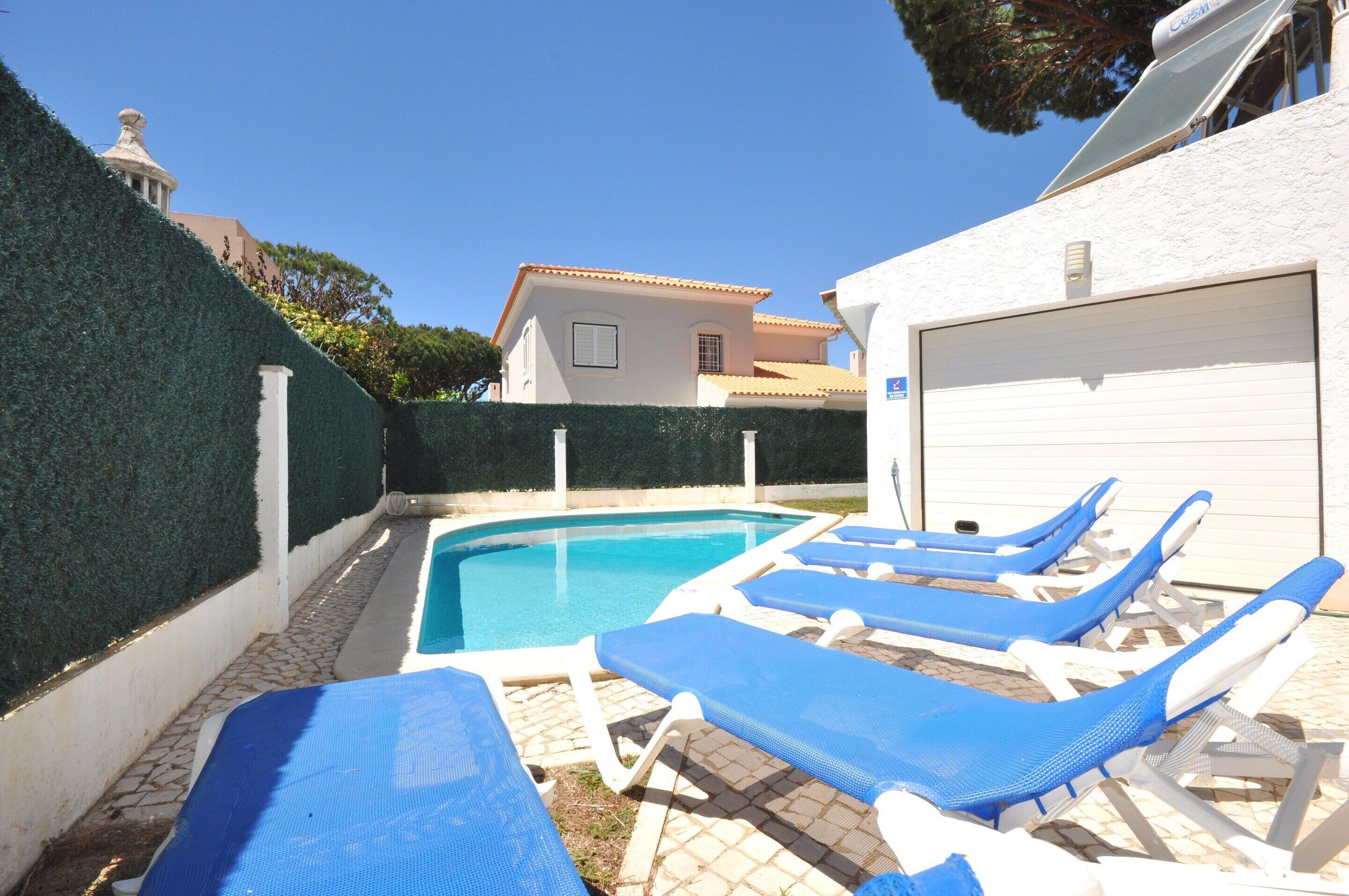 A swimming pool with blue lounge chairs, a white garage, and a building with a tiled roof in the background.