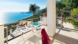 En balkon med pool, spisebord og stole, der giver udsigt over kysten med bjerge.