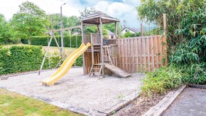 A wooden play structure with a slide and climbing area, surrounded by a fence and greenery. - Pleasant Holiday Home With Infrared Sauna (Durbuy)