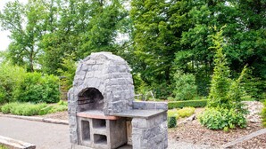 A stone-built outdoor oven structure in a landscaped garden area with surrounding greenery and a paved walkway. - Pleasant Holiday Home With Infrared Sauna (Durbuy)