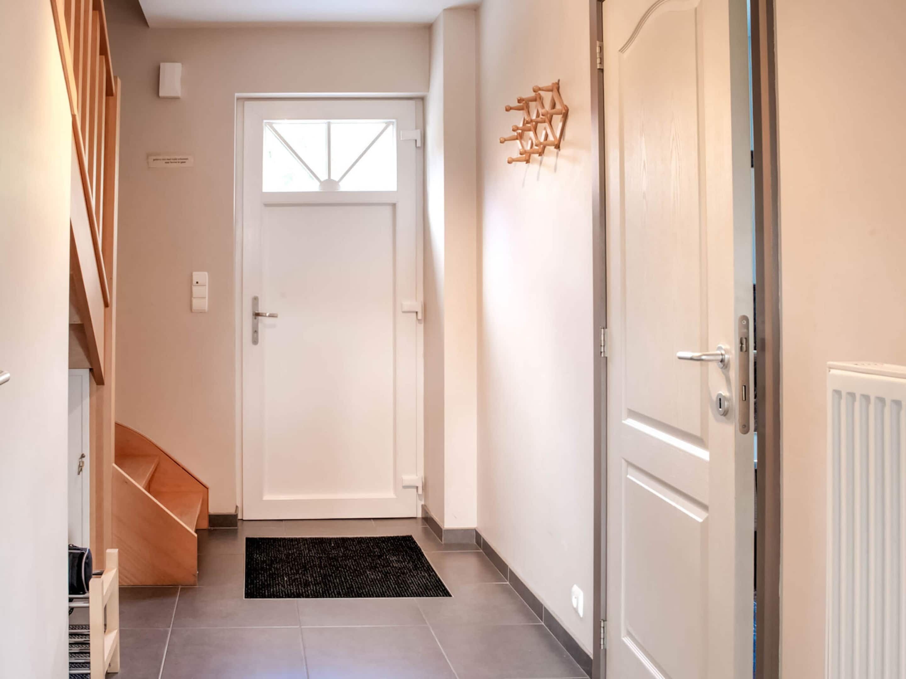 Un corridoio con una porta bianca, un tappetino nero, una scala in legno e un termosifone.