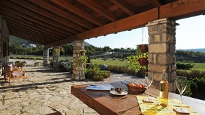 An outdoor patio with stone flooring, wooden furniture, and a table set with a bottle, glasses, and a basket. - Picturesque Mansion in Kozljak (Krsan)