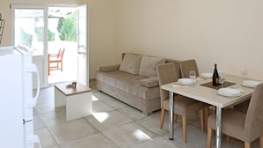 A modern living room with a beige sofa, a dining table set for two, and a kitchen area with a refrigerator and a small table with a plant. - Modern Ground Floor Apartment With Private Terrace Near the Sea (Razanac)