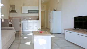 A modern kitchen with white cabinets, a microwave, and a refrigerator. There is a dining area with a table and chairs, and a living area with a sofa and a TV. - Modern Ground Floor Apartment With Private Terrace Near the Sea (Razanac)