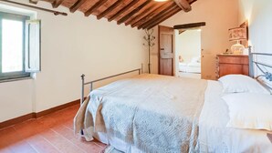 Ein Schlafzimmer mit Holzbalkendecke, einem Bett mit weißer Bettdecke, einem Nachttisch mit Lampe und einem Fenster mit geöffnetem Klappladen.