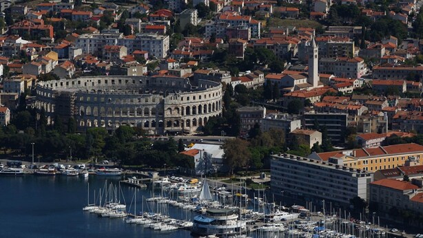 A coastal cityscape with a historic building, a marina with numerous boats, and residential buildings with red-tiled roofs.