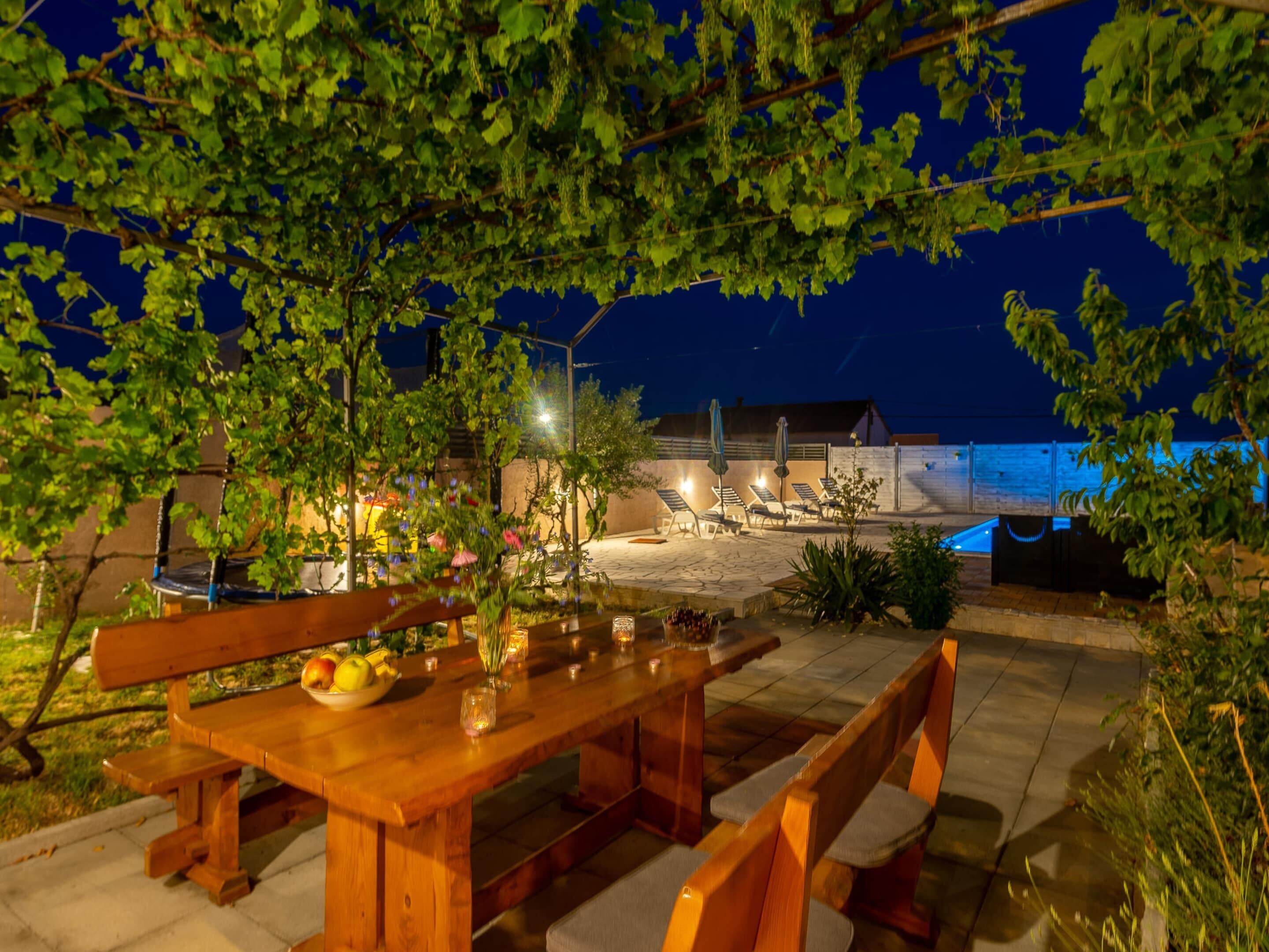 Uma área de refeições ao ar livre com uma mesa e bancos de madeira, rodeada por vegetação e com vista para um edifício ao fundo.