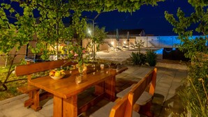 Un área de comedor al aire libre con mesa y bancos de madera, rodeada de follaje y con vista a un edificio al fondo.