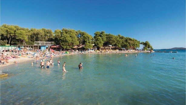 Una playa con agua cristalina, gente nadando y tomando el sol, y una hilera de árboles y edificios a lo largo de la costa.
