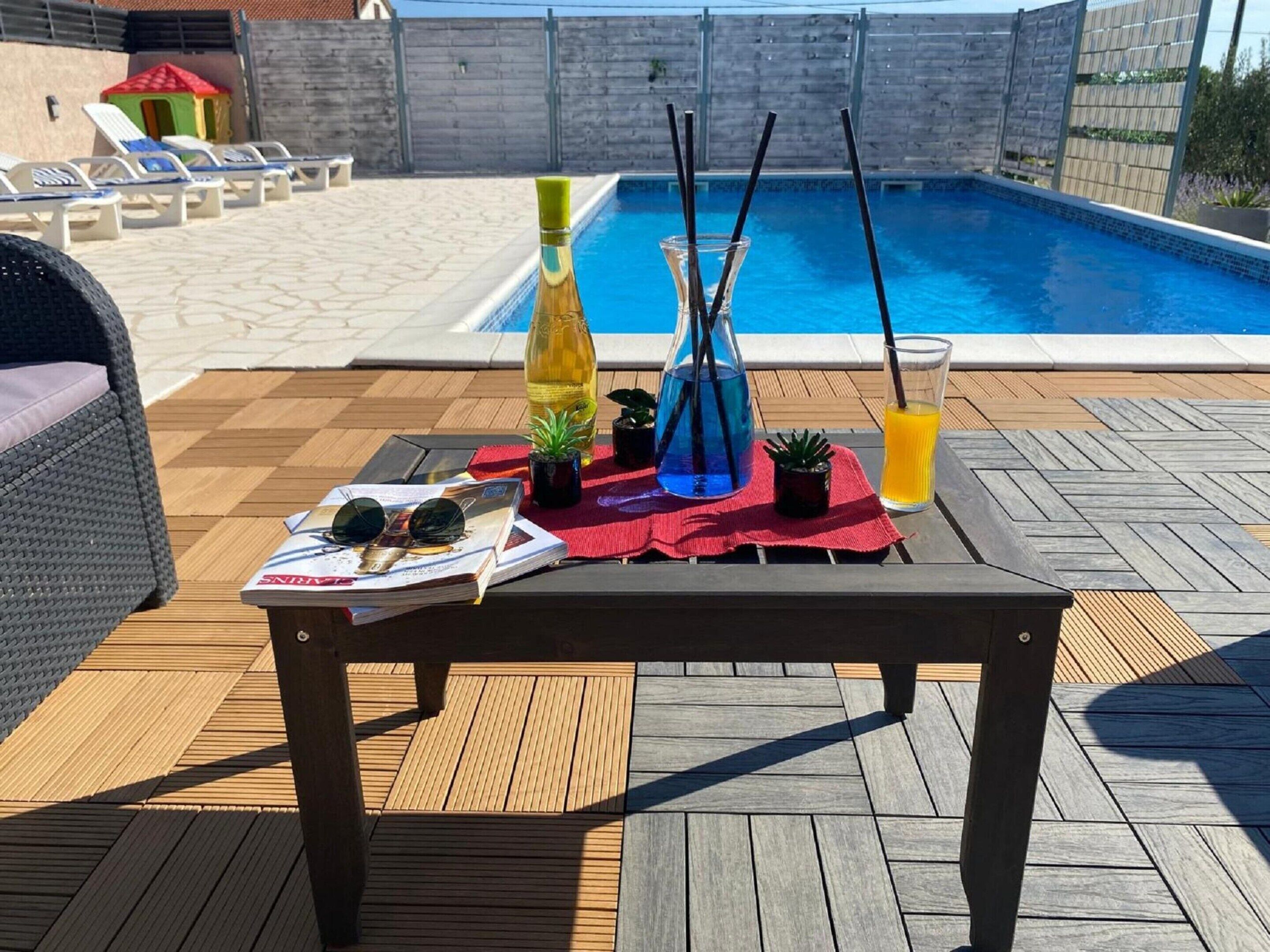 Uma área na piscina com um deck de madeira, espreguiçadeiras, uma mesa com bebidas e uma revista, e a piscina ao fundo.