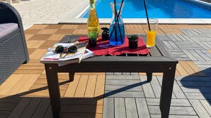 Un área junto a la piscina con una terraza de madera, sillas de descanso, una mesa con bebidas y una revista, y una piscina al fondo.