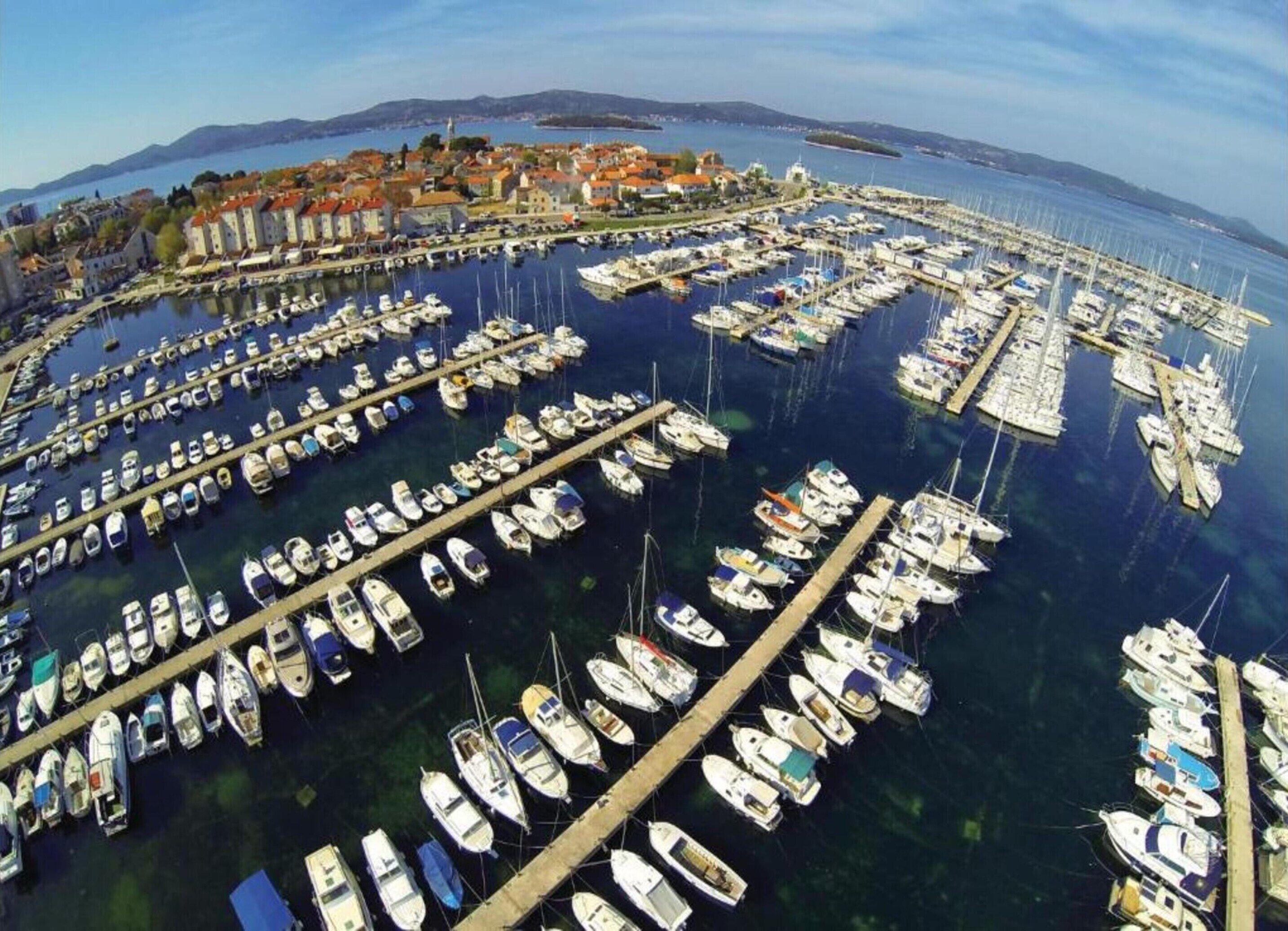 Vista aérea de um porto com inúmeros barcos atracados em um padrão circular, cercado por uma cidade costeira com edifícios e um céu claro.