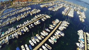 Vista aérea de un puerto deportivo con numerosas embarcaciones amarradas en un patrón circular, rodeado por un pueblo costero con edificios y un cielo despejado.