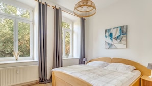 A bedroom with a wooden bed, large windows with curtains, a rattan ceiling light, and a modern abstract painting.