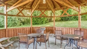 A wooden pavilion with a covered patio area, furnished with metal tables and wicker chairs, and a grill.