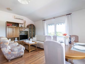 Un salon comprenant un canapé, des fauteuils, une table basse et une table à manger pour deux personnes. On y trouve également un téléviseur, des étagères et une grande fenêtre avec des rideaux.