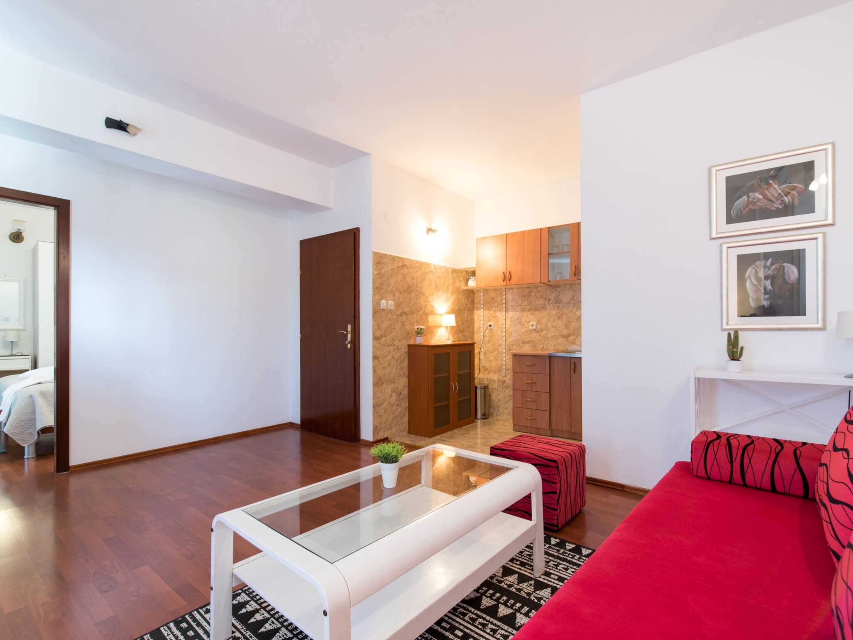 A modern living room with a glass coffee table, a red sofa, and a kitchen area with wooden cabinets and a marble backsplash.