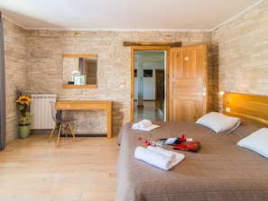Un dormitorio con pared de piedra, un escritorio de madera, una cama con almohadas y una mesita de noche con un libro y una botella.