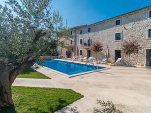 Un edificio de piedra con piscina, mobiliario de exterior y un gran árbol.