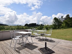 An outdoor patio with a table and chairs, a decorative metal stand, and a view of a grassy field and trees under a partly cloudy sky. - Holiday Home in Leglise Near Ardennes River (Léglise)