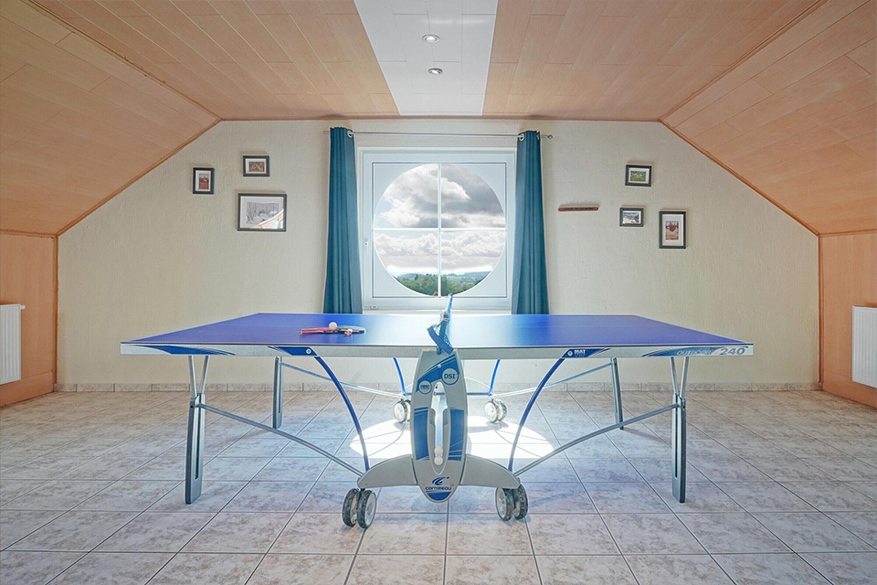 Une table de ping-pong se trouve dans une pièce dont le plafond est en pente, avec une grande fenêtre munie de rideaux et plusieurs tableaux encadrés accrochés aux murs.