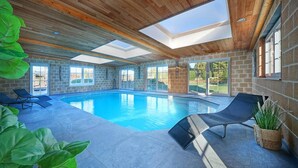Una amplia piscina interior con techo de madera y tragaluces, rodeada de sillones de descanso y plantas en macetas.