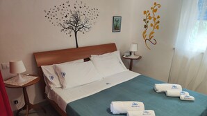 A neatly made bed with a teal bedspread and white pillows, a small bedside table with a lamp, a wall clock, and a decorative tree decal. - Apartment in Alcamo Near the Beach (Alcamo)