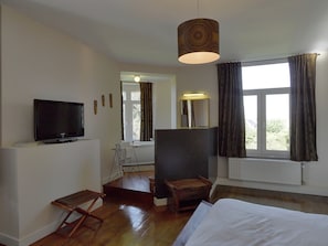 A hotel room with a bed, a television, a window with curtains, a mirror, and a small wooden stool.