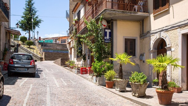 Una calle adoquinada flanqueada por edificios, un coche aparcado y macetas con plantas.