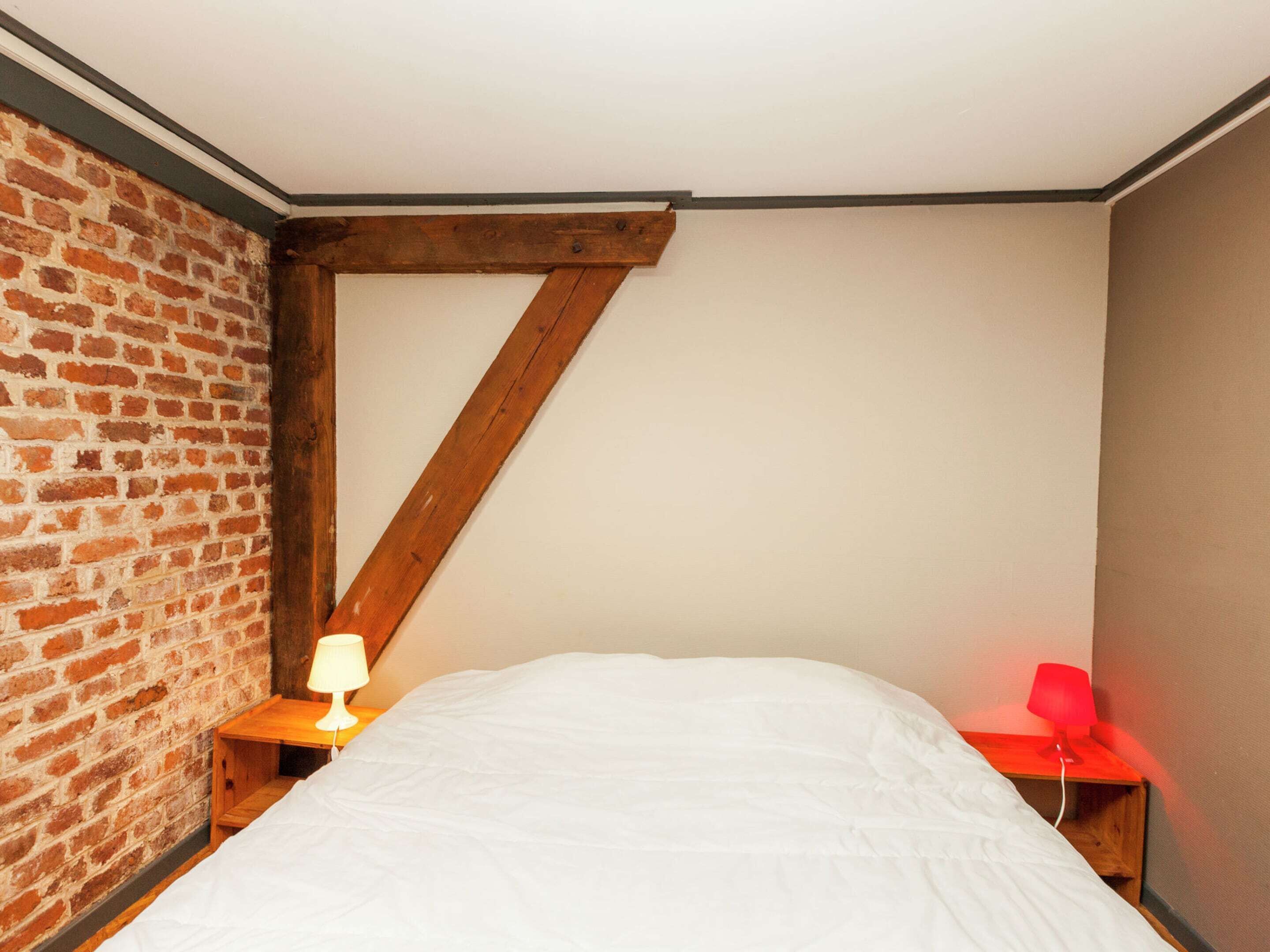 Um quarto com parede de tijolos, teto com vigas de madeira, cama com roupa de cama branca, mesa de cabeceira com luminária e abajur vermelho no criado-mudo.