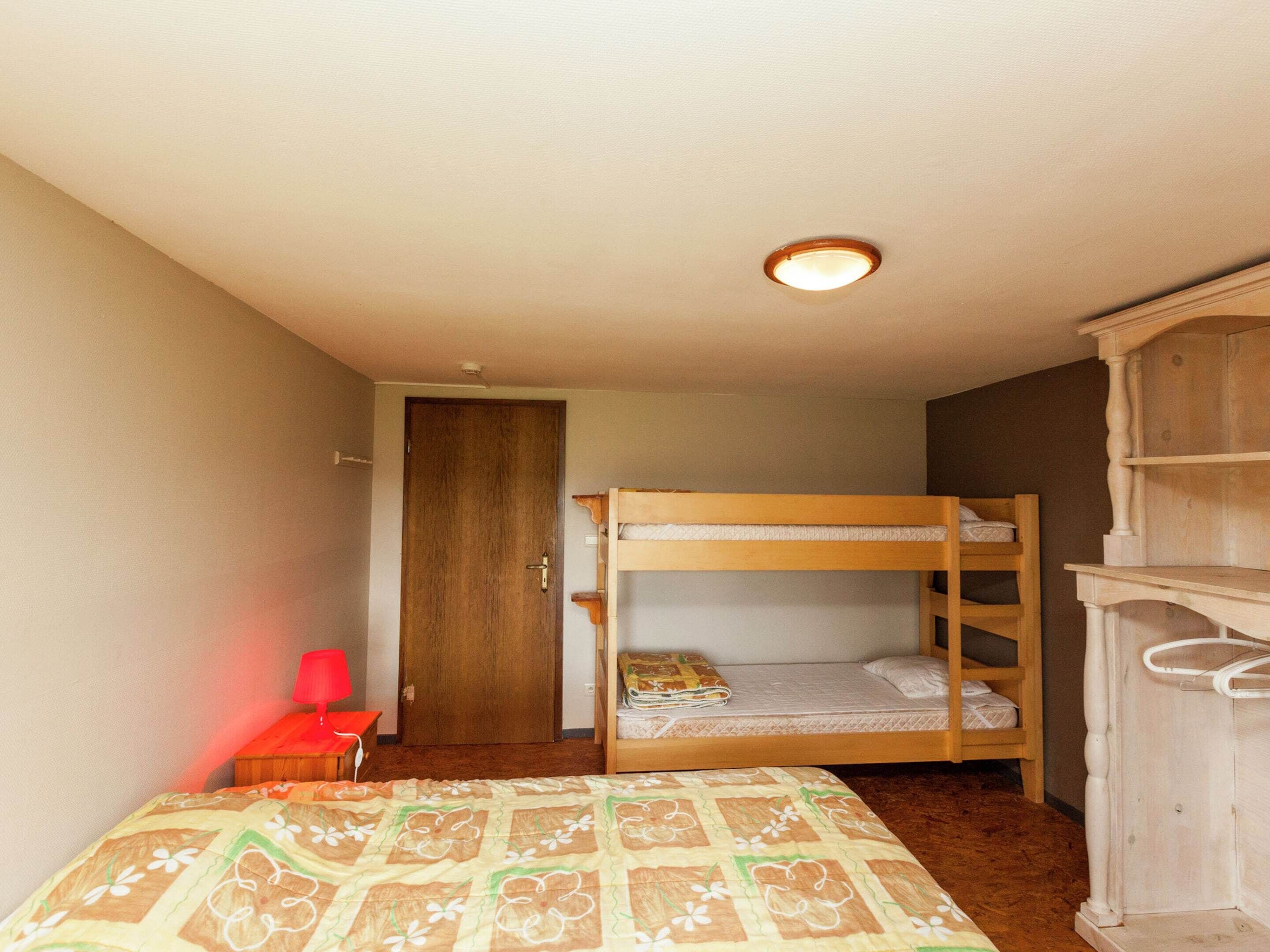 Habitació petita i senzilla amb un llit superposat, un armari de fusta, una cadira vermella i una taula de nit.