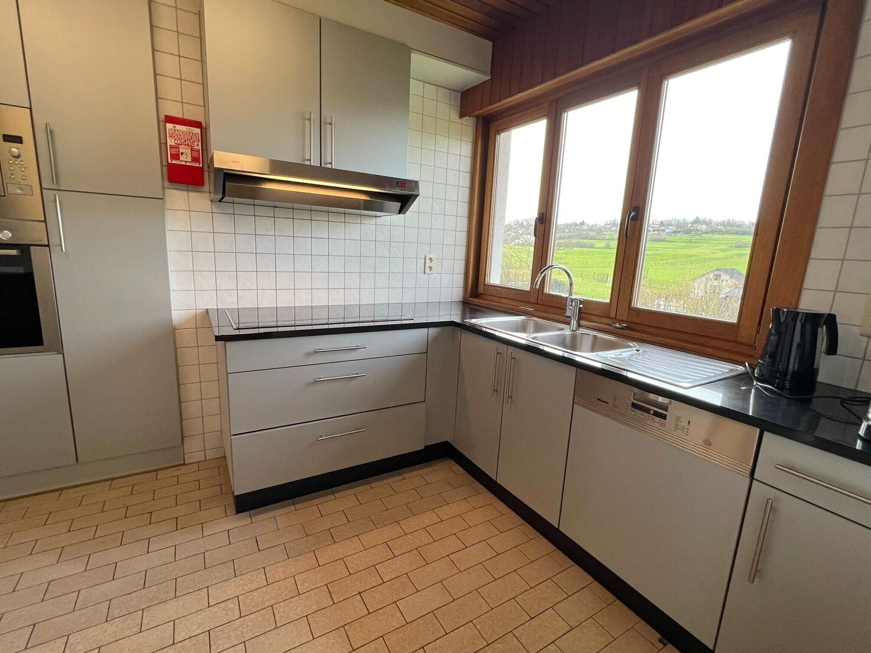 A kitchen with white cabinets, a stainless steel range hood, a double sink, and a window with a view of a green field.