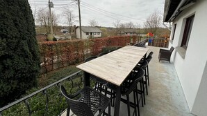 A balcony with a wooden table and black chairs, overlooking a residential area with houses and a hedge. - House in Noiseux With Bubble Bath & Garden (Somme-Leuze)