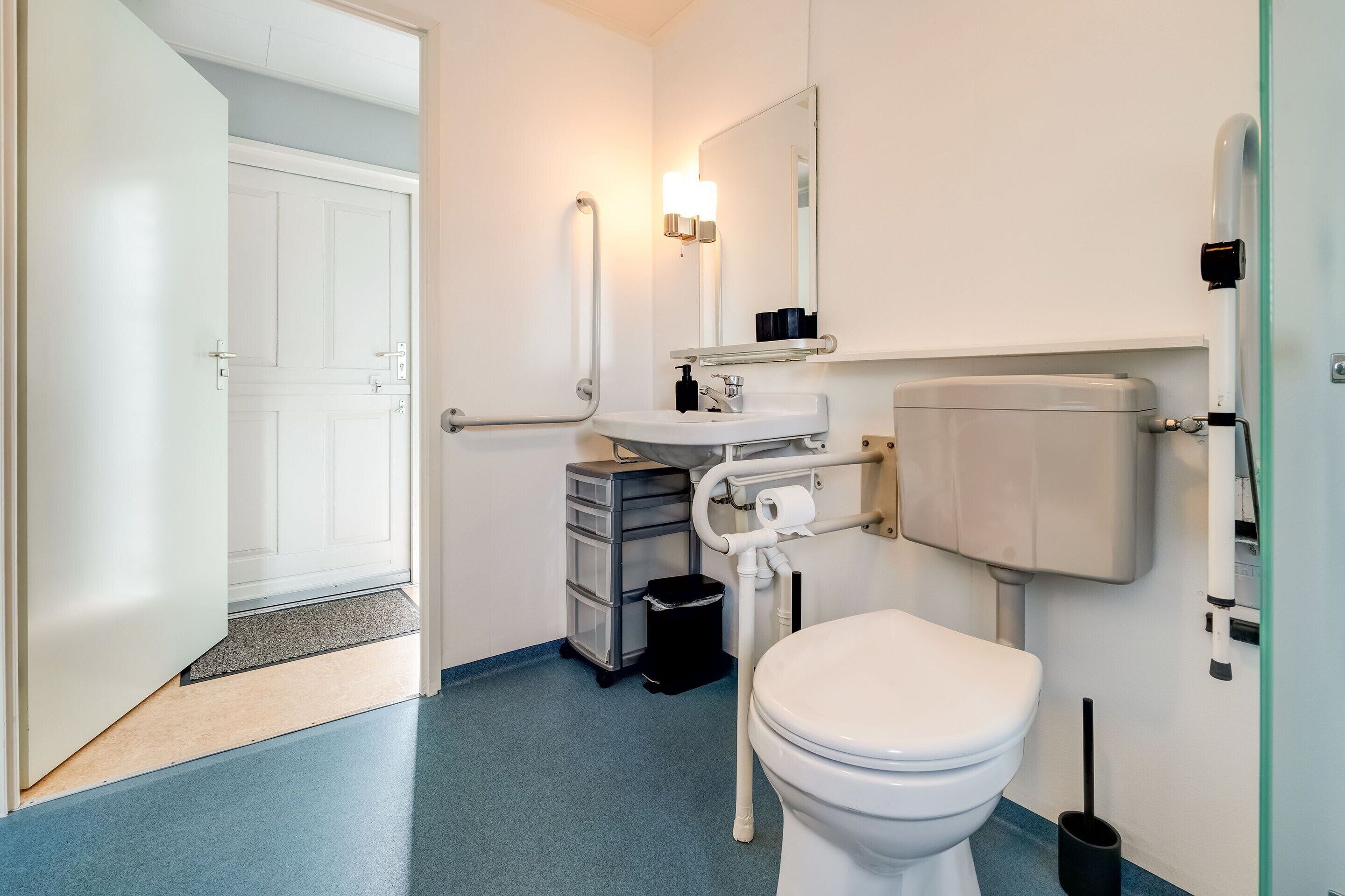 A modern bathroom with a white toilet, a sink, and a mirror. There is a door leading to another room and a trash bin next to the sink.
